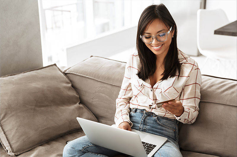 asian businesslike woman 20s holding credit card and using laptop, while sitting at sofa in apartment
